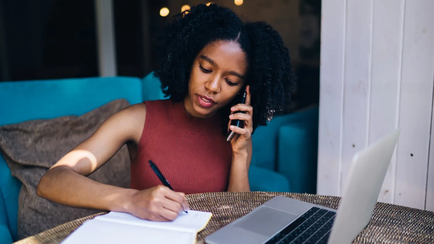 A woman on a phone call, writing notes beside her laptop