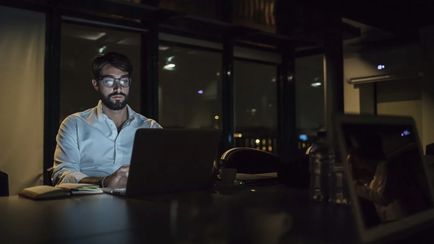 A man working late at a laptop, alone in a dark office